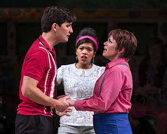 Orlando in his red with stripes polo shirt and Orlando as Ganymede sans jacket hold both hands as the face each other. In the back is Celia in a simple gray patterned blouse.
